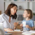 Happy child and adult are sitting at desk. Girl doing homework or online education.